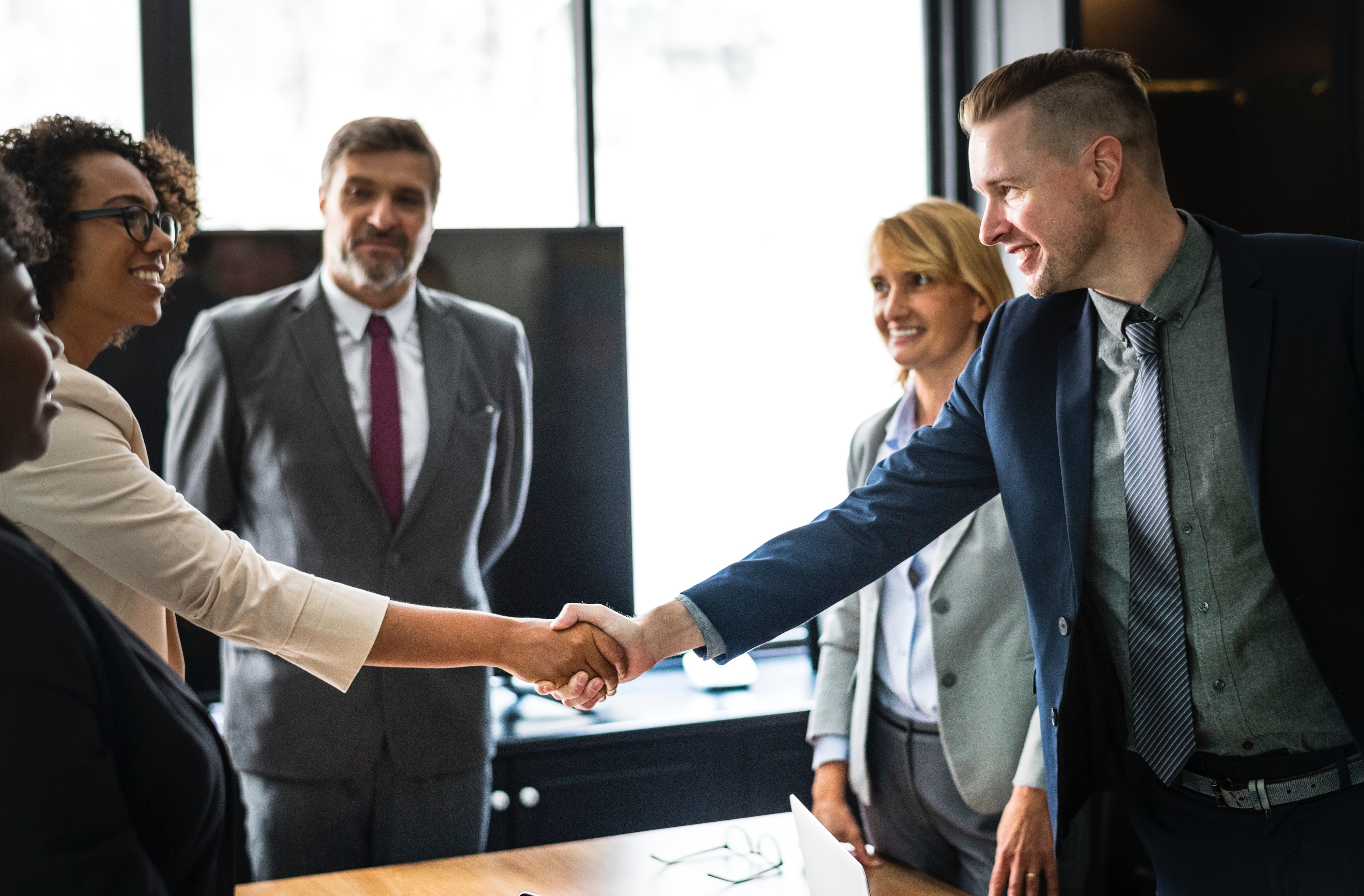 businessmen-businesspeople-businesswomen-job interview A PR job interview candidate in a suit and tie shaking hands with a hiring manager dressed in a white suit, with other interviewers watching